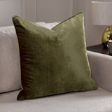Luxe Velvet Plain Polyester Cushion in olive green, square with piped edges, shown on a light beige sofa beside a table lamp.