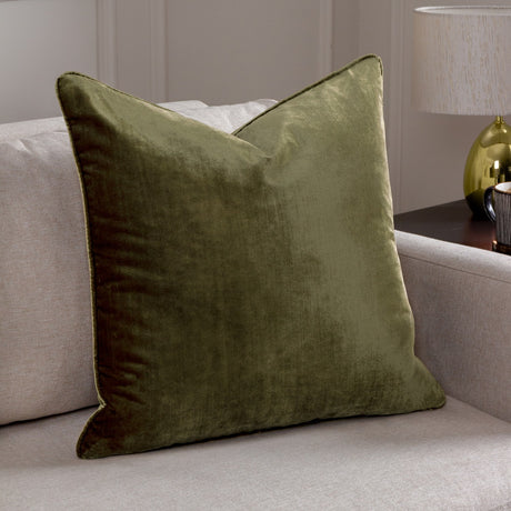 Luxe Velvet Plain Polyester Cushion in olive green, square with piped edges, shown on a light beige sofa beside a table lamp.