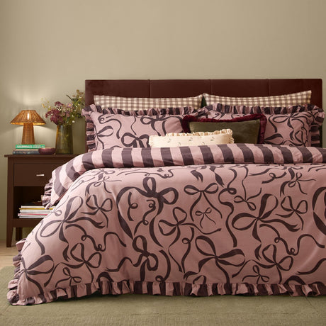 Bedroom scene with La Vie En Bows reversible duvet cover set in mocha-pink, featuring aubergine bow print and striped reverse, with ruffled edges and matching pillowcases on a dark headboard bed.