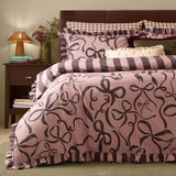 La Vie En Bows reversible duvet cover set in mocha-pink with aubergine bow print, frilled trim, and striped reverse, shown styled on a bed with matching pillowcases.
