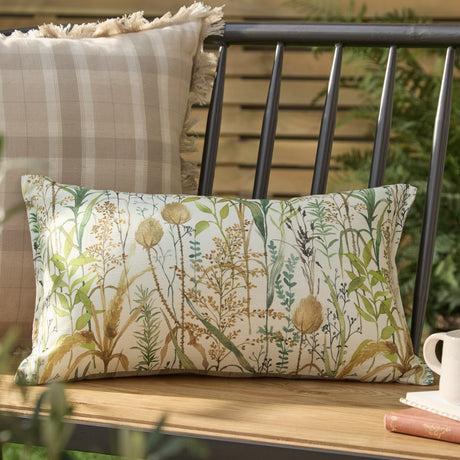 Lydiard linen cotton bolster cushion in Moss, rectangular with a watercolour meadow print in green, amber and brown tones, shown on an outdoor bench beside a check cushion.