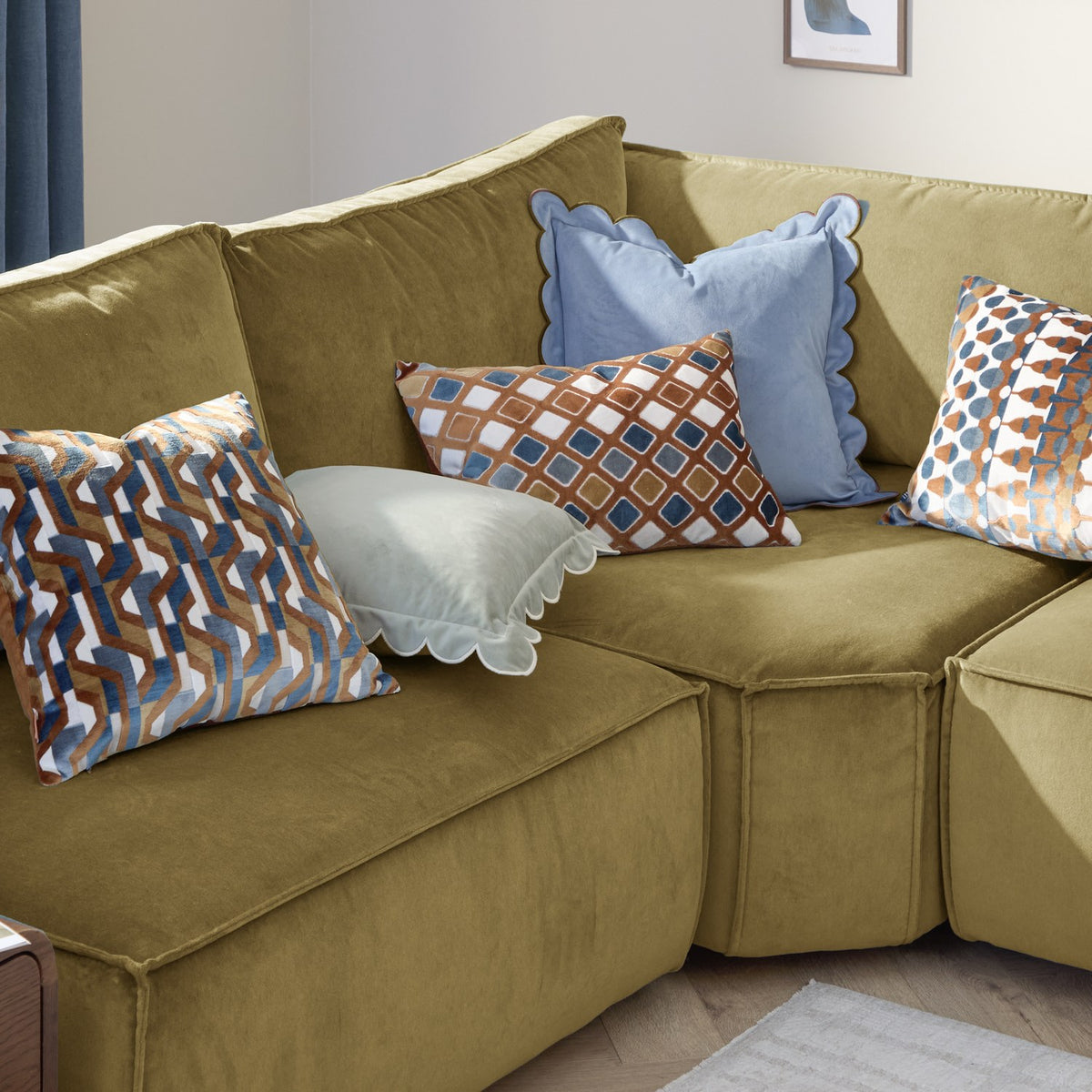 Metro Abstract Jacquard Velvet Cushion in Ginger with rust, teal and denim geometric pattern on cream, shown on an olive green sofa among coordinating patterned and plain cushions.