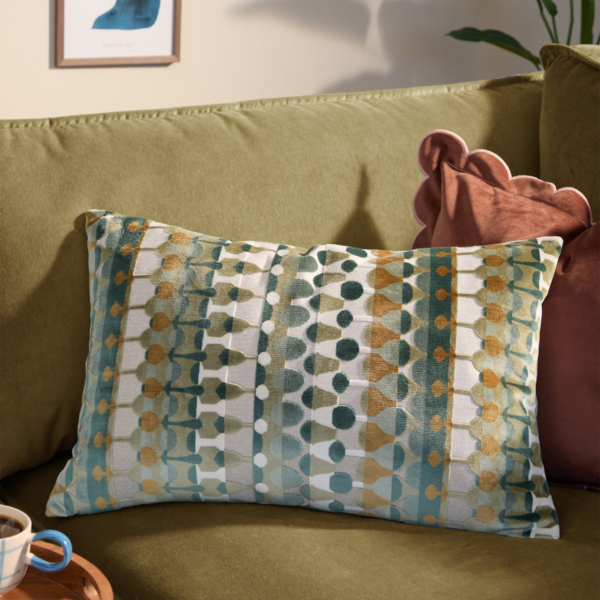Metro Abstract Jacquard Velvet Cushion in multicoloured green, teal and amber geometric pattern, shown on an olive-green sofa beside a rust pink cushion.