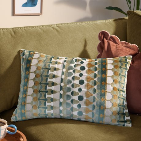 Metro Abstract Jacquard Velvet Cushion in multicoloured green, teal and amber geometric pattern, shown on an olive-green sofa beside a rust pink cushion.
