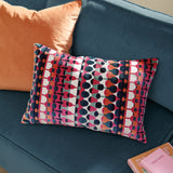 Metro Abstract Jacquard Velvet Cushion in multicoloured geometric pattern with pink, orange, teal and navy stripes, shown on a blue velvet sofa beside an orange cushion.