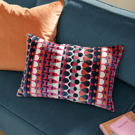 Metro Abstract Jacquard Velvet Cushion in multicoloured geometric pattern with pink, orange, teal and navy stripes, shown on a blue velvet sofa beside an orange cushion.