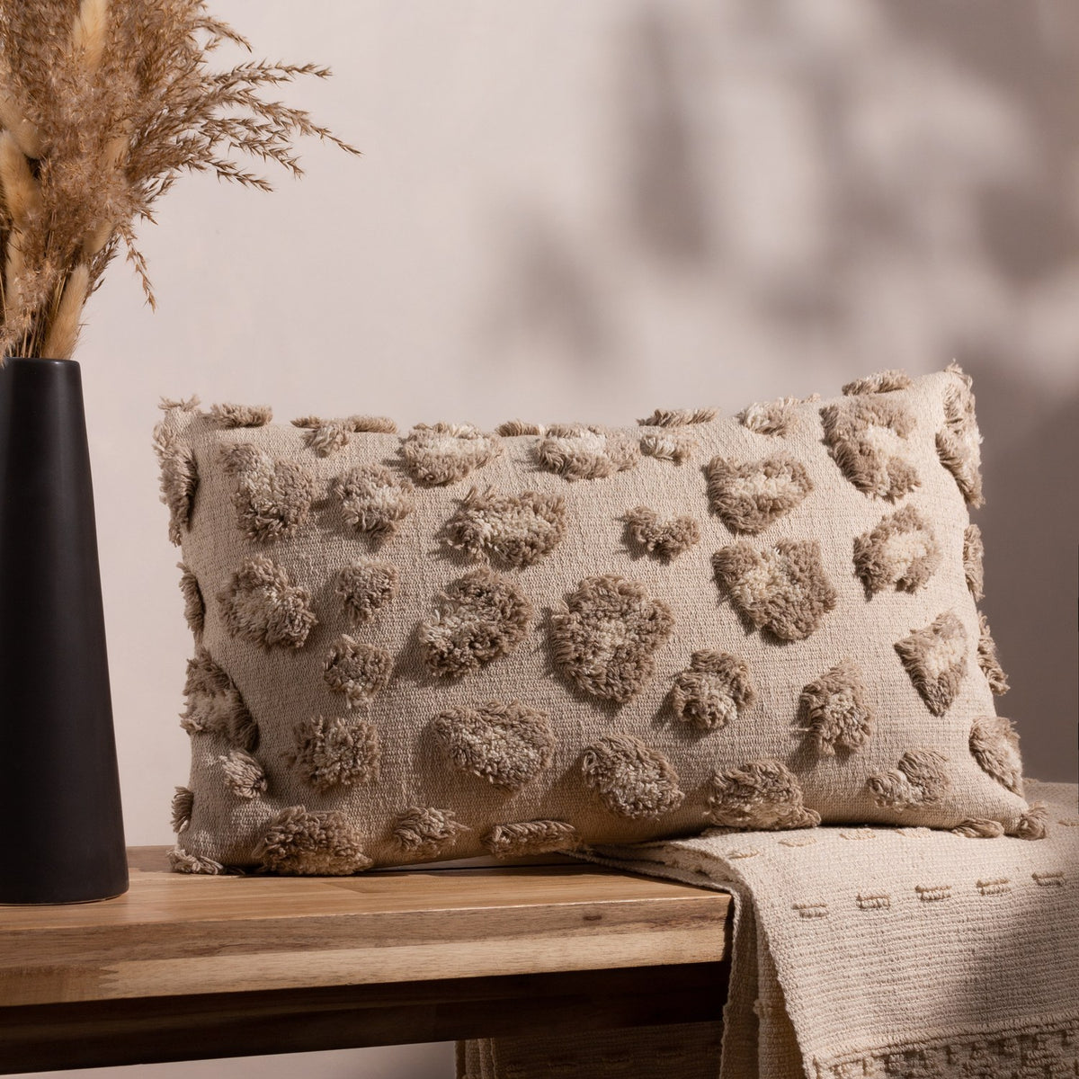 Maeve Animal Cotton Cushion in natural, a rectangular bolster with raised tonal leopard-print tufts, shown on a wooden bench beside dried grasses and a woven throw.