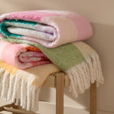 Maisie Check Polyester Throw folded on a stool with pink, teal, lilac and cream check pattern, fringed edges, shown atop other multicoloured throws.