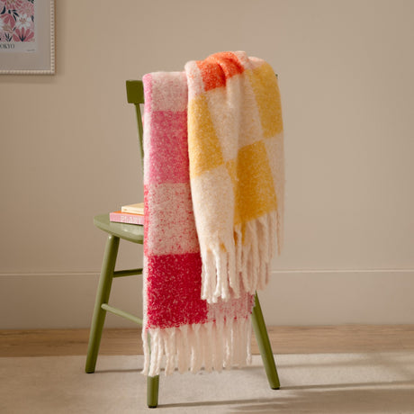 Maisie Check Polyester Throw in Citrus Blush, a fringed woven blanket in cream, yellow, orange and pink checks, draped over a green wooden chair.