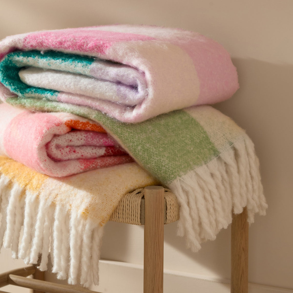 Maisie Check Polyester Throw in Citrus Blush, folded with pink, teal and coral check patterns visible, stacked with other throws on a wooden stool.
