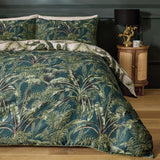 Majorelle Cotton Leaf Duvet Cover Set in green, styled on a bed with matching pillowcases showing a tropical palm and fern print, in a bedroom with a wooden bedside table and lamp.