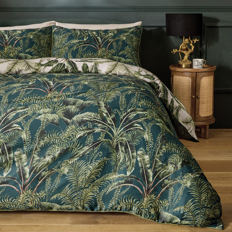 Majorelle Cotton Leaf Duvet Cover Set in green, styled on a bed with matching pillowcases showing a tropical palm and fern print, in a bedroom with a wooden bedside table and lamp.