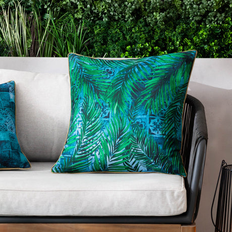 Majorelle Reversible Polyester Cushion in Azure, square outdoor cushion with green palm leaf print and caramel piping, shown on a beige sofa against a leafy garden backdrop.