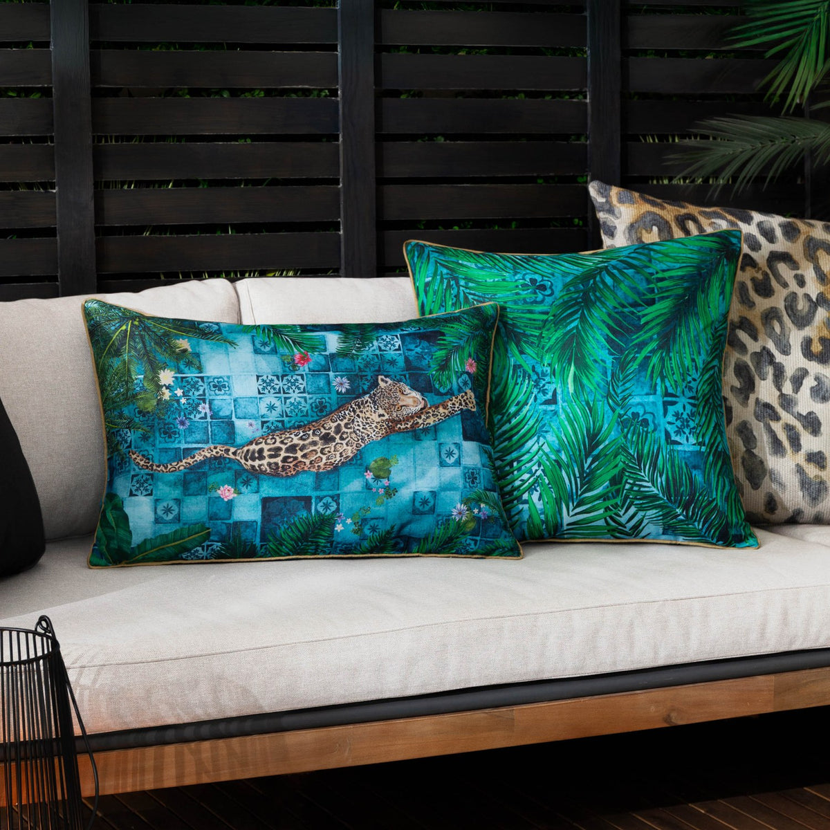 Majorelle Reversible Polyester Cushion in Azure, square with green palm leaf print and caramel piping, styled on a cream sofa with leopard-print and tile-motif cushions.