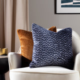Malans Geometric Velvet Cushion in bronze with cut-velvet crackled pattern, shown on a cream armchair beside a navy patterned cushion in a living room setting.