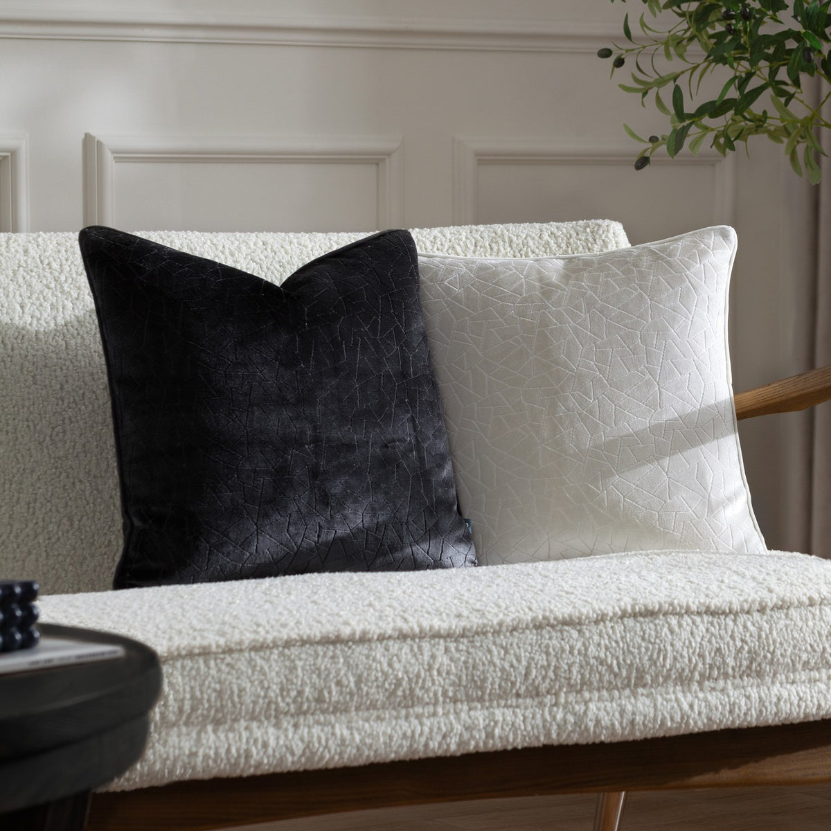 Malans Geometric Velvet Cushion in milk cream with a crackled-glass textured pattern, shown beside a black version on a cream boucle sofa.