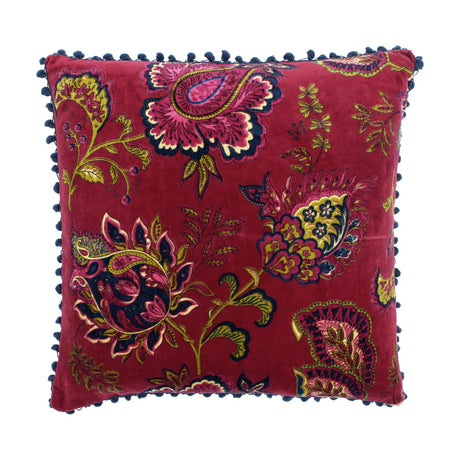 Malisa Velvet Cotton Cushion in Pomegranate, a square red cushion with paisley and floral print in pink, gold-green and blue, edged with blue pompom trim on a white background.
