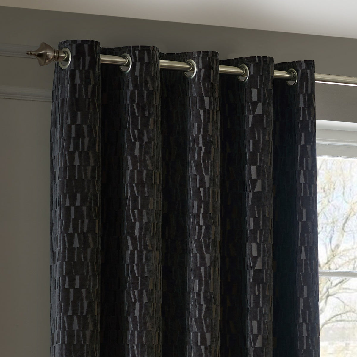 Manhattan Velvet Fully Lined Eyelet Curtains in charcoal, showing eyelet headings on a chrome pole with a textured geometric pile, hung by a window in a living room.