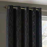 Manhattan Velvet Fully Lined Eyelet Curtains in charcoal, showing eyelet headings on a chrome pole with a textured geometric pile, hung by a window in a living room.