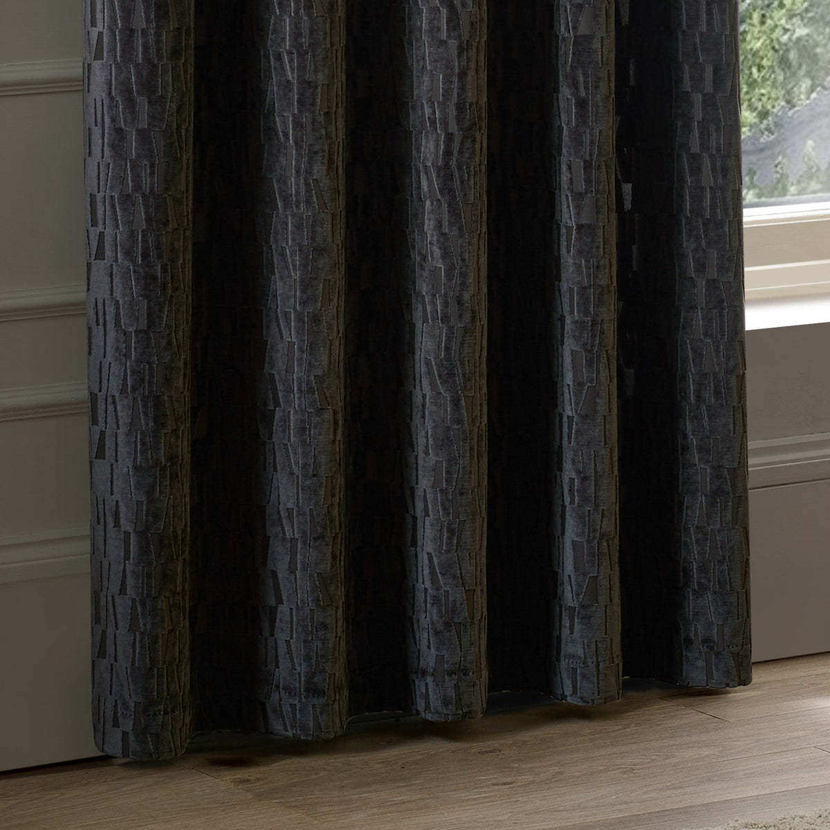 Manhattan Velvet Fully Lined Eyelet Curtains in charcoal, showing textured geometric cut-pile fabric with eyelet heading, draped floor-length beside a window in a living room setting.