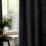 Manhattan Velvet Fully Lined Eyelet Curtains in charcoal, textured cut-velvet fabric with vertical geometric facets, shown hanging beside a window in a living room setting.