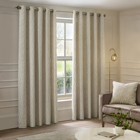 Manhattan fully lined eyelet curtains in ivory cut velvet with a textured geometric pattern, shown on brass rings framing a window in a living room.