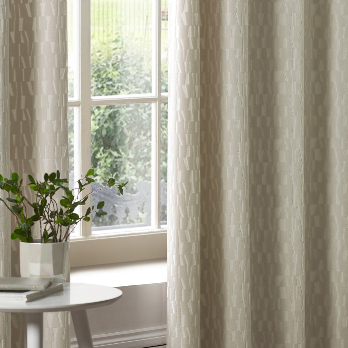Manhattan fully lined eyelet curtains in ivory cut velvet with a textured geometric block pattern, shown hanging beside a window in a living room setting.