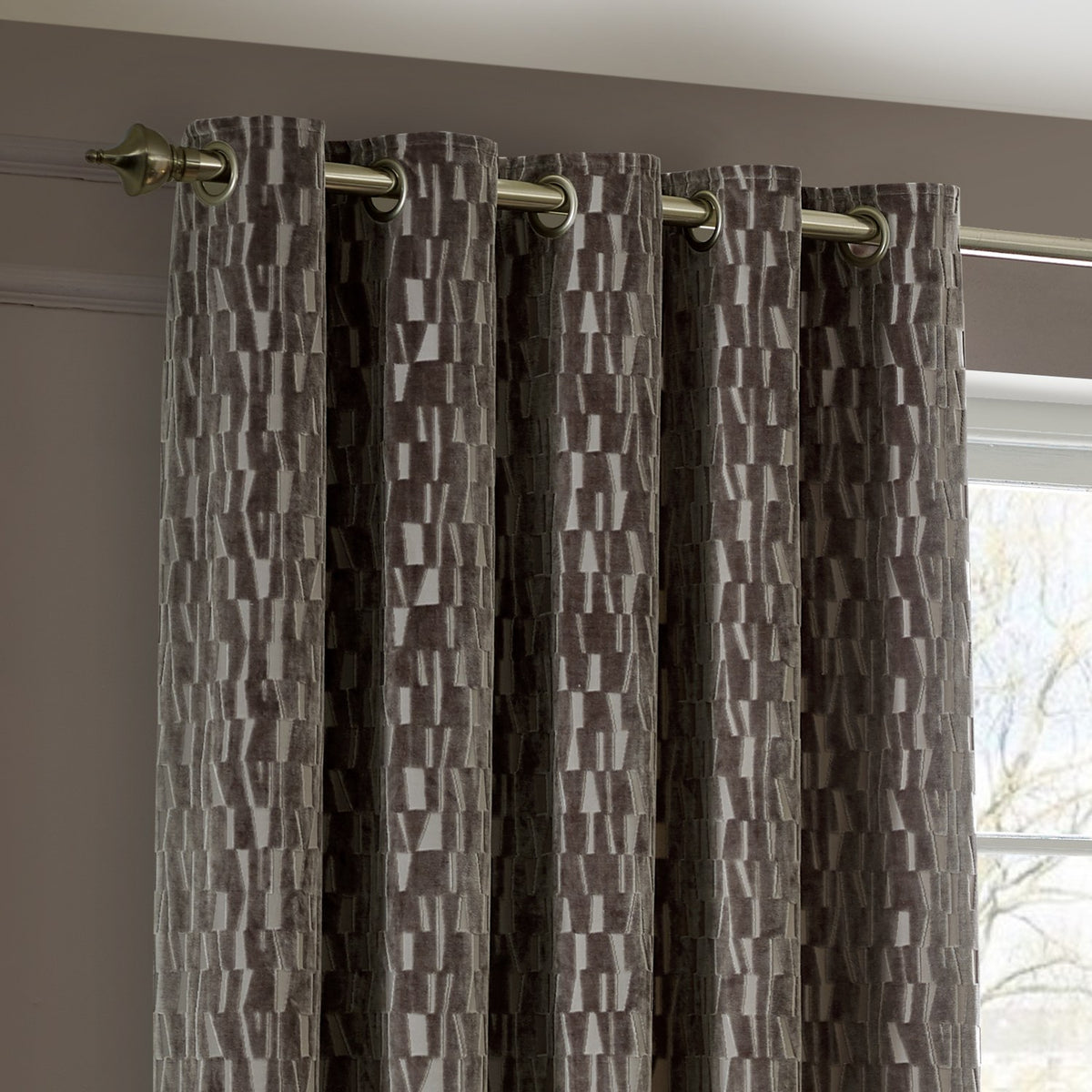 Manhattan Velvet Fully Lined Eyelet Curtains in mink brown with a textured geometric cut-velvet pattern, brass eyelets on a metal pole in a living room setting.