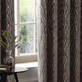 Manhattan Velvet Fully Lined Eyelet Curtains in mink brown with textured geometric pattern, shown hanging by a window in a living room setting.