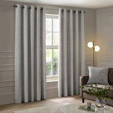 Manhattan Velvet Fully Lined Eyelet Curtains in silver grey with a raised geometric pattern, shown either side of a window on a metal pole in a living room setting.