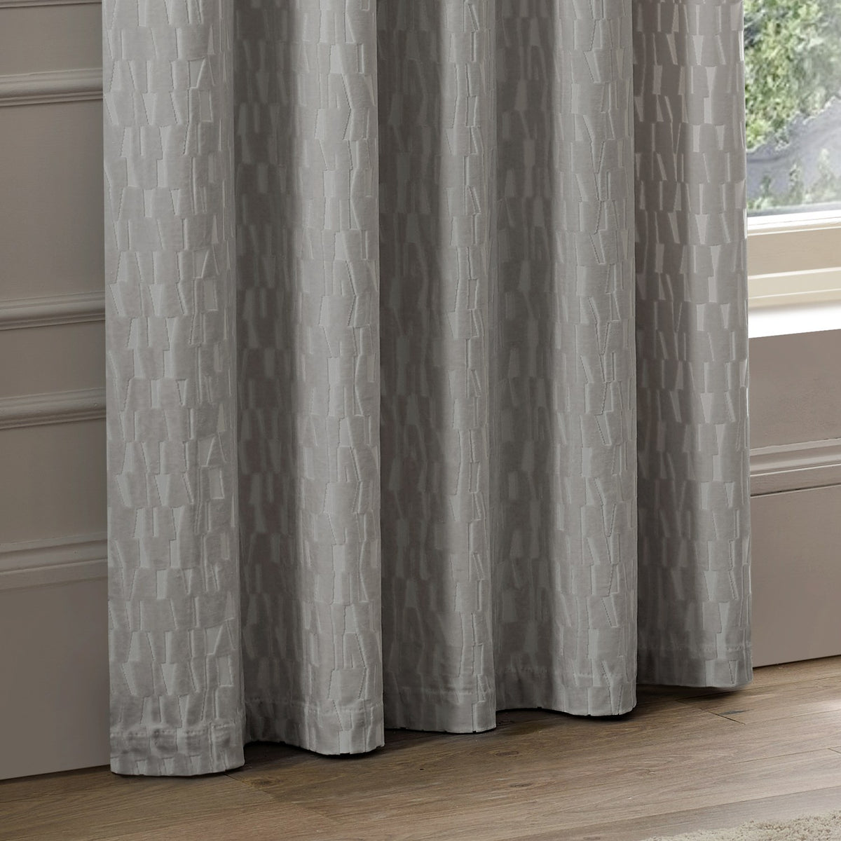 Manhattan Velvet Fully Lined Eyelet Curtains in silver grey, shown with soft folds and a raised geometric block pattern, hanging beside a window in a living room setting.