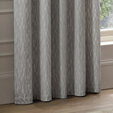 Manhattan Velvet Fully Lined Eyelet Curtains in silver grey, shown with soft folds and a raised geometric block pattern, hanging beside a window in a living room setting.