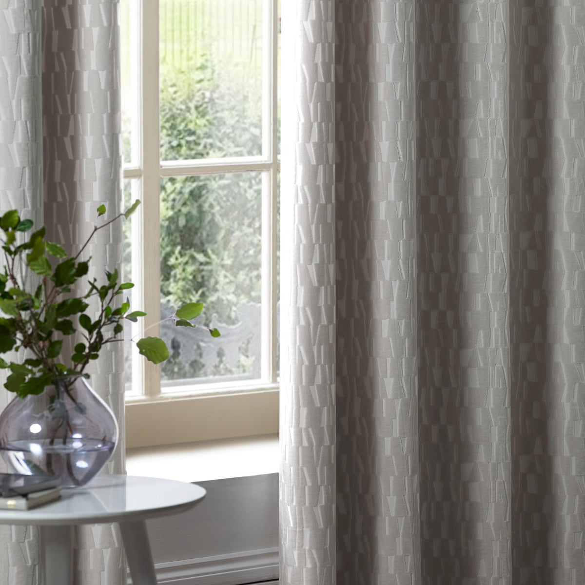 Manhattan Velvet Fully Lined Eyelet Curtains in silver grey, shown hanging by a window with a raised geometric textured pattern in a living room setting.