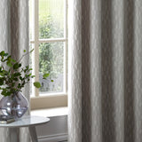 Manhattan Velvet Fully Lined Eyelet Curtains in silver grey, shown hanging by a window with a raised geometric textured pattern in a living room setting.