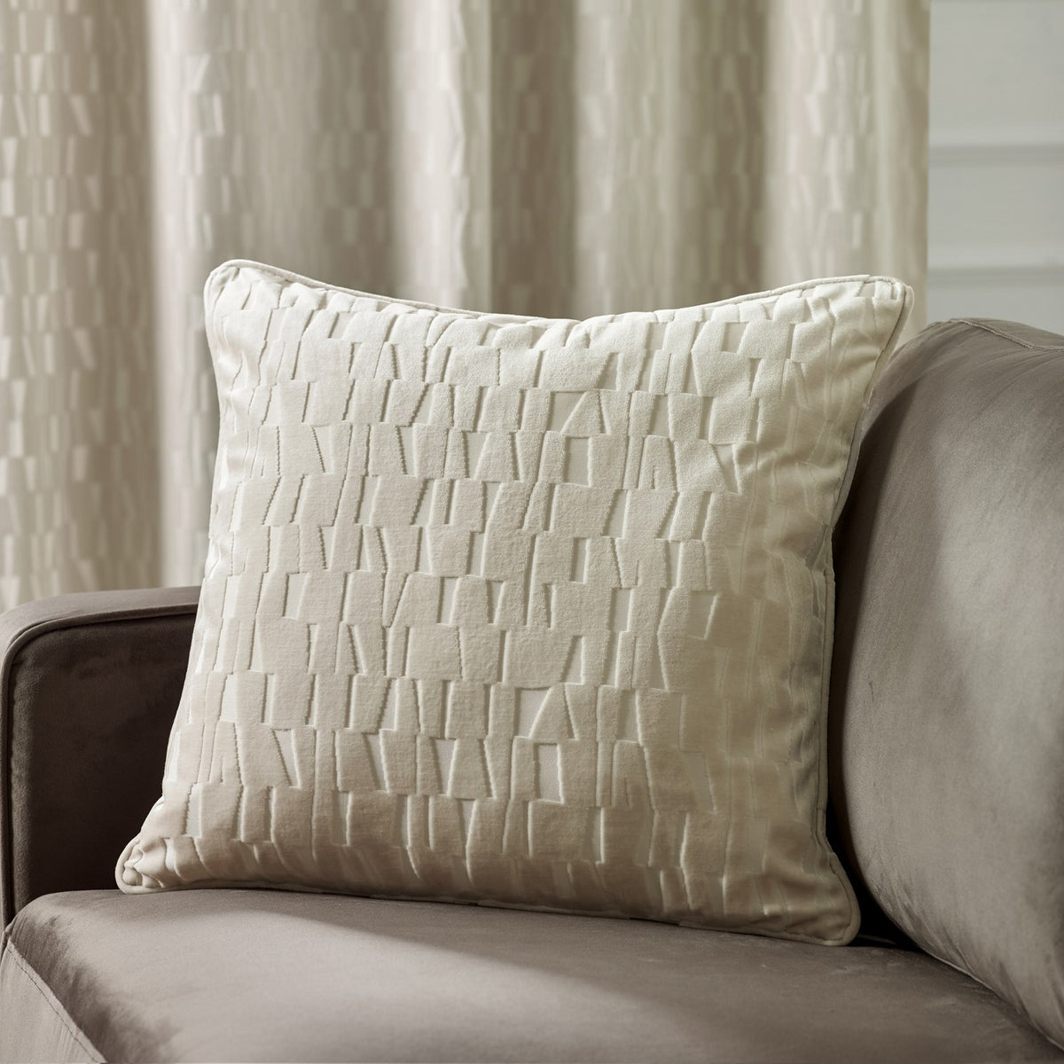 Manhattan Geometric Velvet Cushion in ivory with a raised cut-velvet brickwork pattern, square shape with piped edges, shown on a beige sofa near curtains.