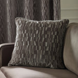 Manhattan Geometric Velvet Cushion in mink with cut-velvet staggered block pattern, shown on a beige sofa against a grey curtain backdrop.