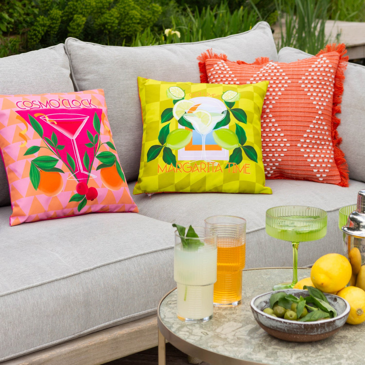 Square polyester cushion in lime-green checked print with a margarita cocktail graphic, limes and "Margarita Time" text, styled on a grey outdoor sofa.