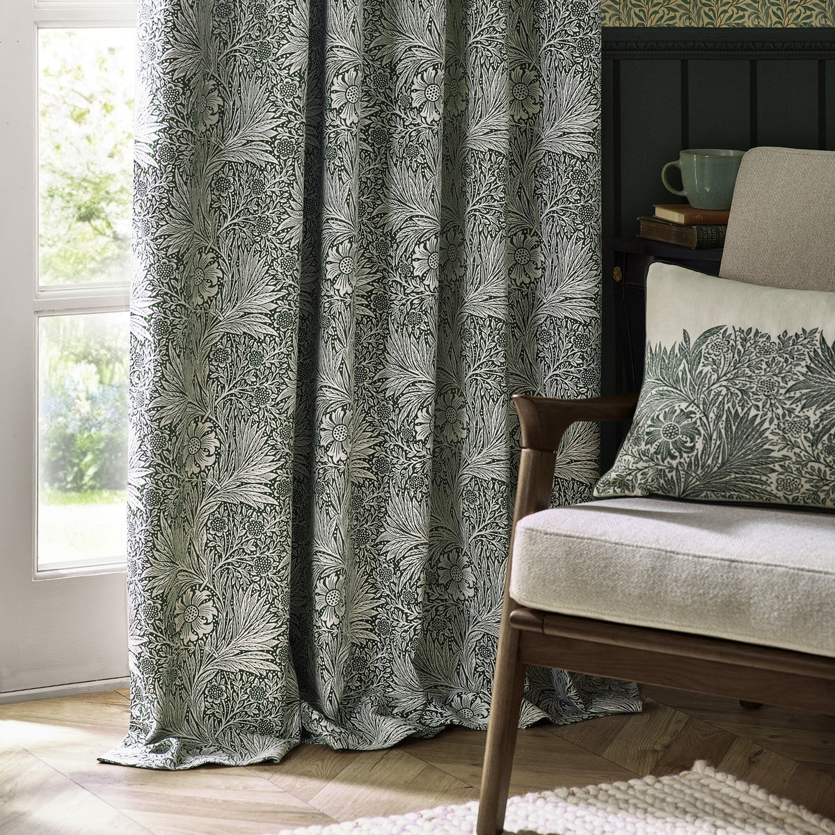 Pencil pleat curtains in leaf green Marigold jacquard, shown pooling by a window beside an armchair with a matching cushion in a living room setting.