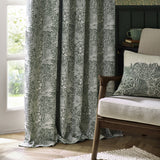 Pencil pleat curtains in leaf green Marigold jacquard, shown pooling by a window beside an armchair with a matching cushion in a living room setting.