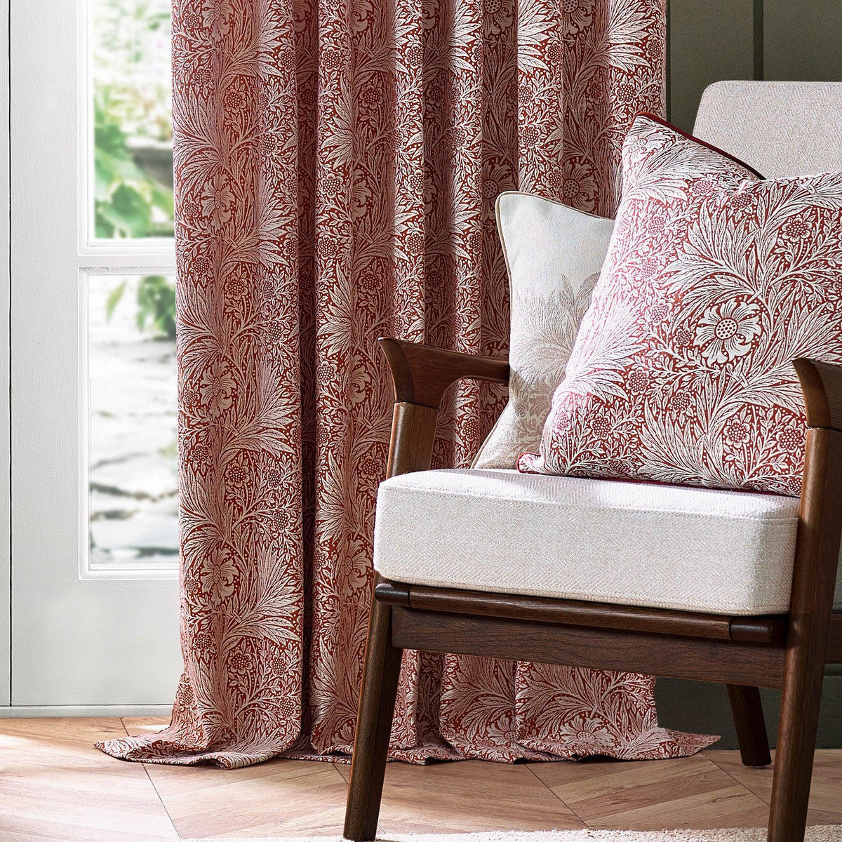 Marigold Jacquard pencil pleat curtains in russet with woven foliage and floral pattern, shown pooling by a window beside a wooden armchair with matching cushions.