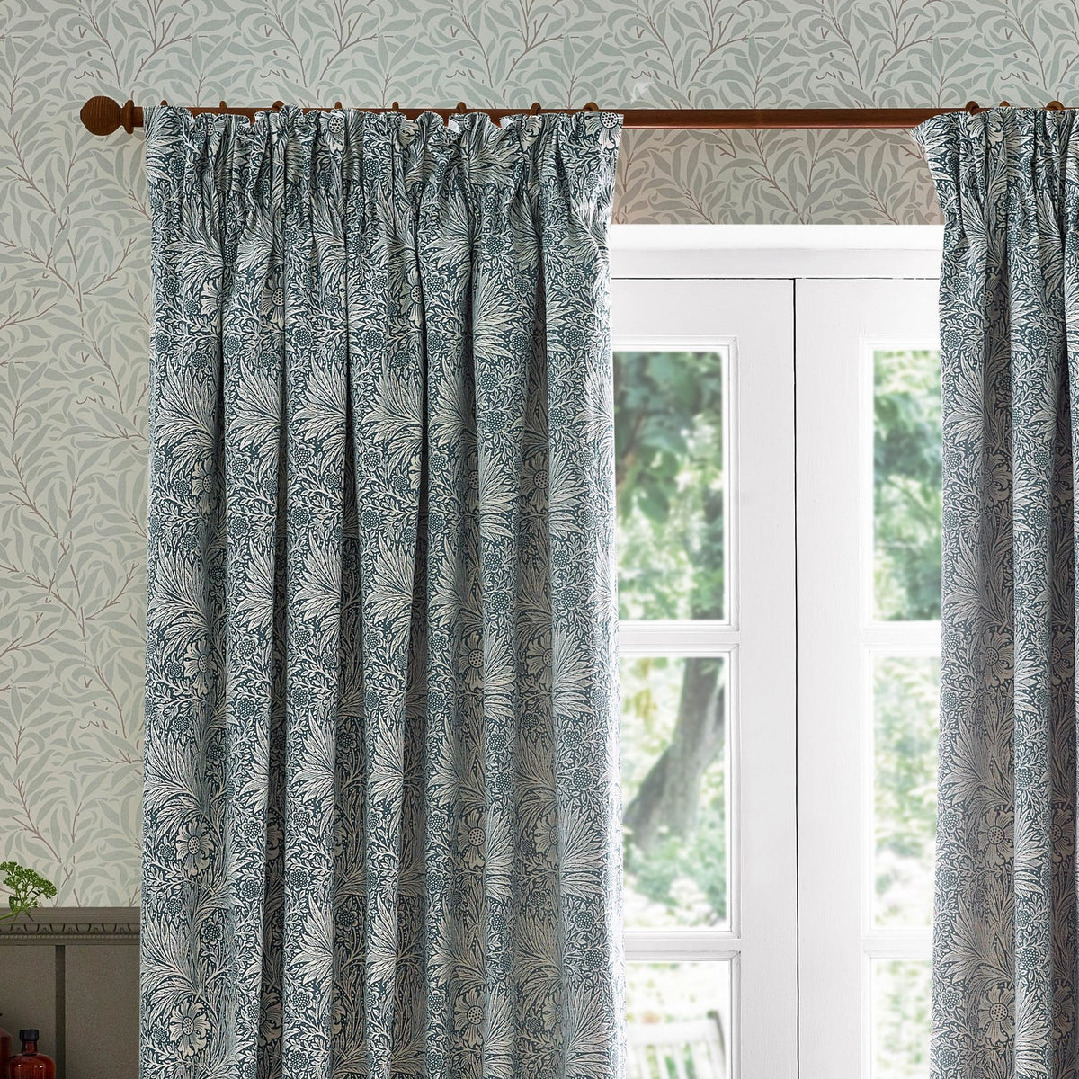 Marigold Jacquard pencil pleat curtains in indigo blue and cream, hung beside a white French door with foliage-patterned wallpaper in the background.