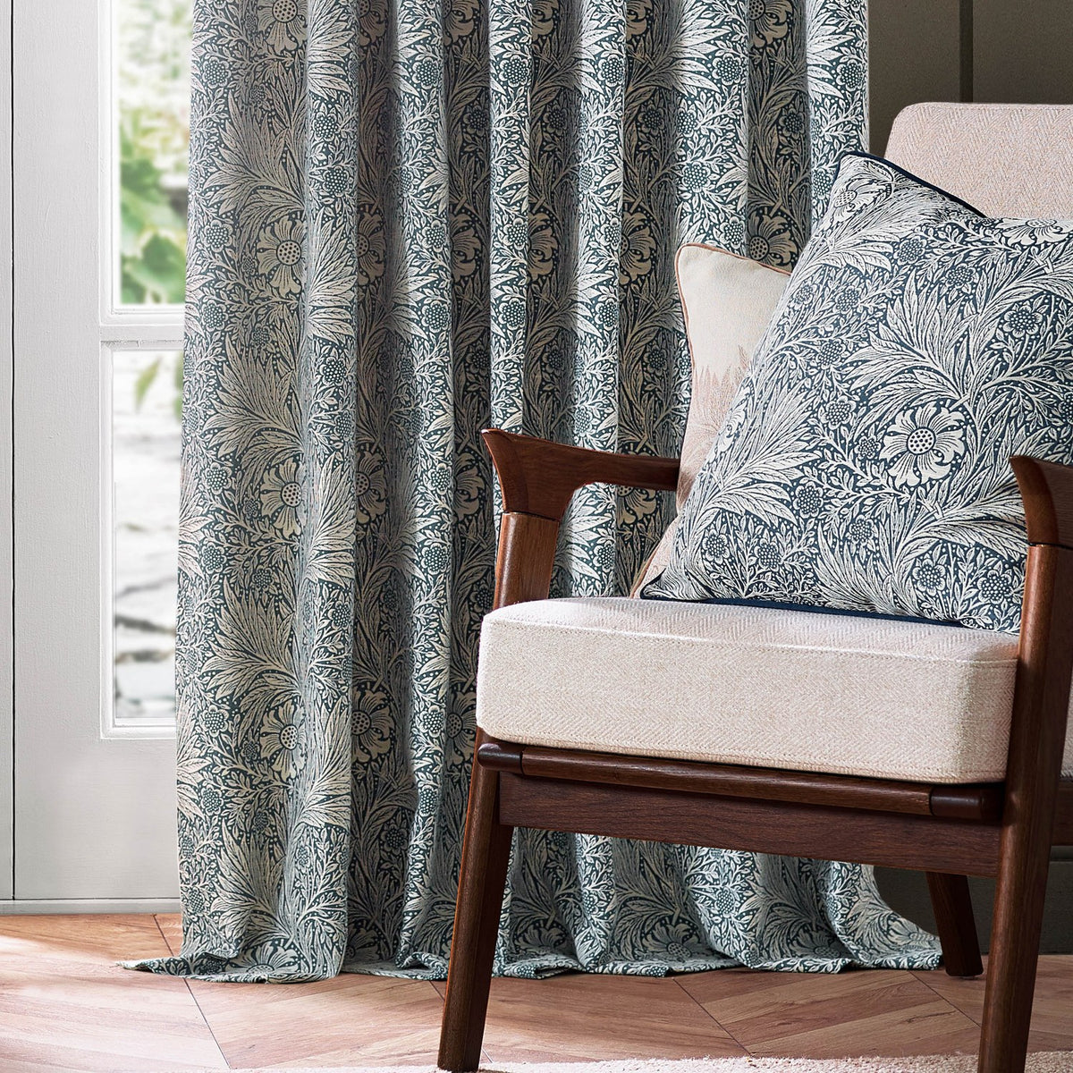 Marigold Jacquard pencil pleat curtains in indigo blue with cream floral pattern, shown pleated beside a window with a matching cushion on a wooden armchair.