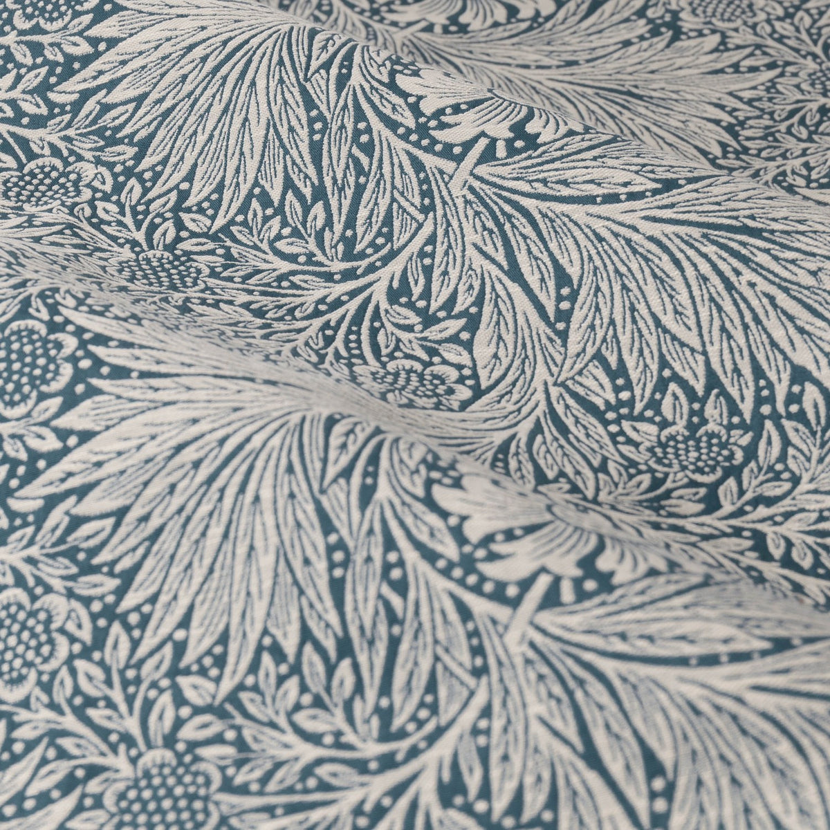 Close-up of Marigold Jacquard pencil pleat curtain fabric in indigo blue, showing swirling foliage and sunburst marigold patterns on a soft cream polyester ground.