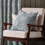 Marigold Jacquard Velvet Cushion in indigo blue with cream floral pattern, shown on a beige armchair with a wooden frame in a living room setting.