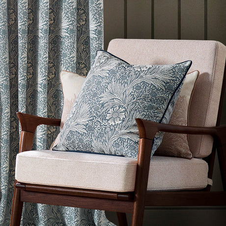 Marigold Jacquard Velvet Cushion in indigo blue with cream floral pattern, shown on a beige armchair with a wooden frame in a living room setting.