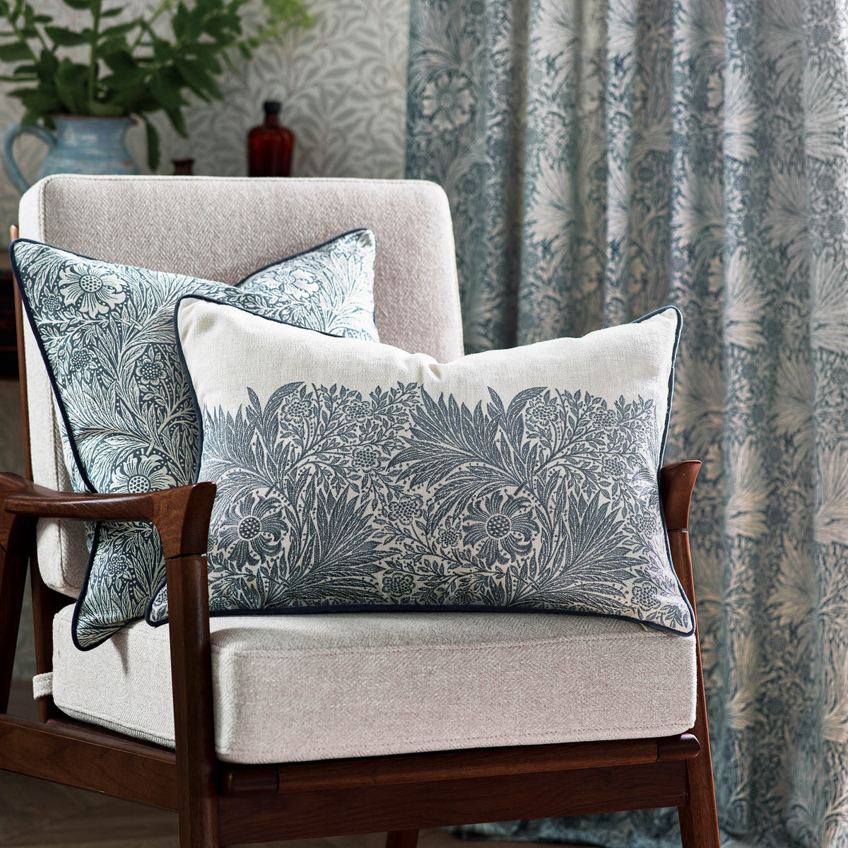 Rectangular Marigold cushion with cream floral jacquard pattern on ivory linen, navy piped edges, styled on a beige armchair with matching patterned curtains.