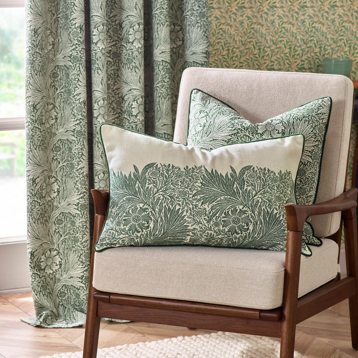 Rectangular Marigold Jacquard Velvet cushion in leaf green with cream marigold pattern and green piping, styled on a beige armchair beside a matching floral cushion and curtains.