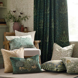 Square stone jacquard velvet cushion with woven Marigold foliage design, shown among coordinating cushions in a green botanical-themed living room setting.