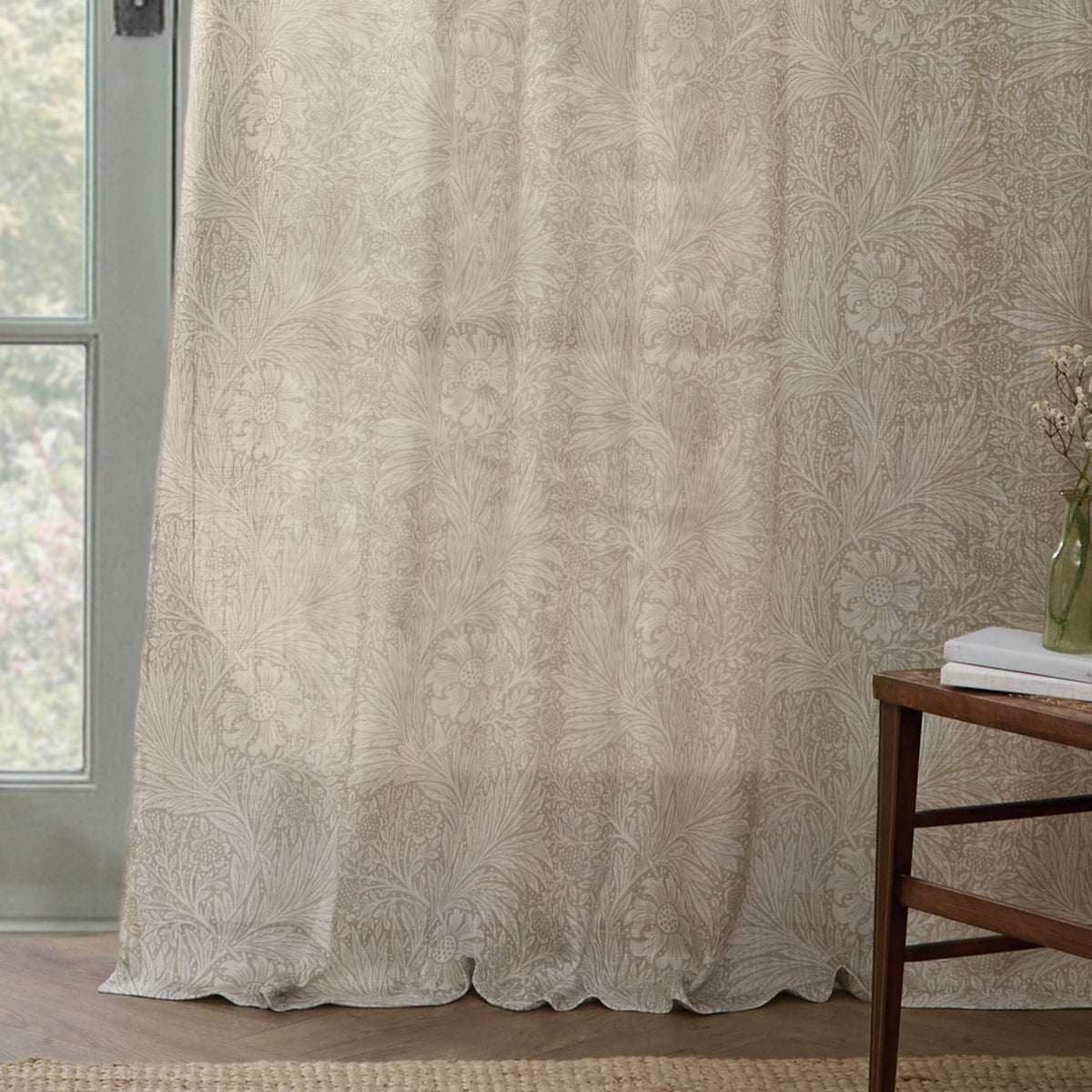 Marigold Damask Linen Curtain Panel in cream, showing a Morris foliage and flower print in putty tone, hung by a glass door beside a wooden side table.