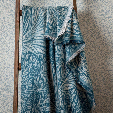 Marigold Jacquard Cotton Throw in indigo, draped over a wooden ladder, showing its woven floral and leaf pattern with a fringed edge against a leaf-print wallpaper backdrop.